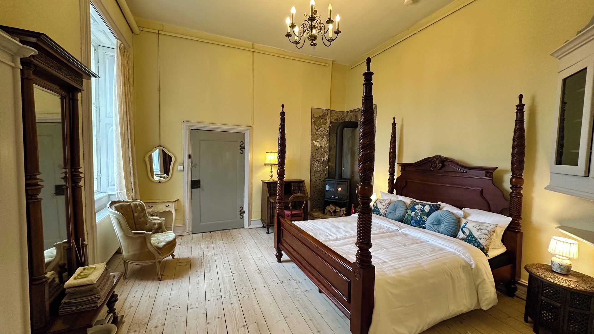 A four-poster bed with a wooden headboard, a sitting chair, a bedside table with a lamp, and a large mirror on the wall.