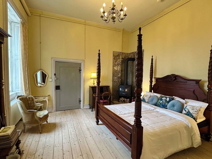 A four-poster bed with a wooden headboard, a sitting chair, a bedside table with a lamp, and a large mirror on the wall.