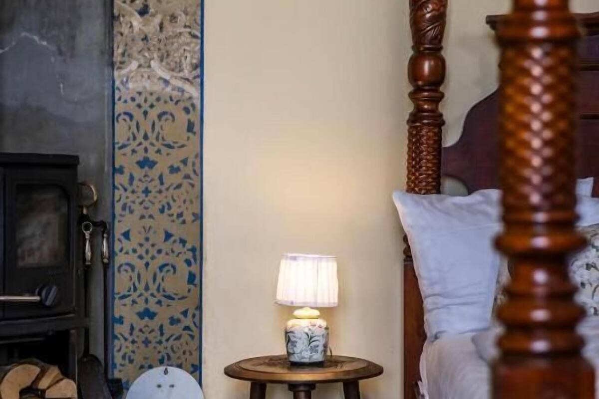 A bedroom with a four-poster bed, a bedside table with a lamp, a fireplace, and patterned wallpaper.