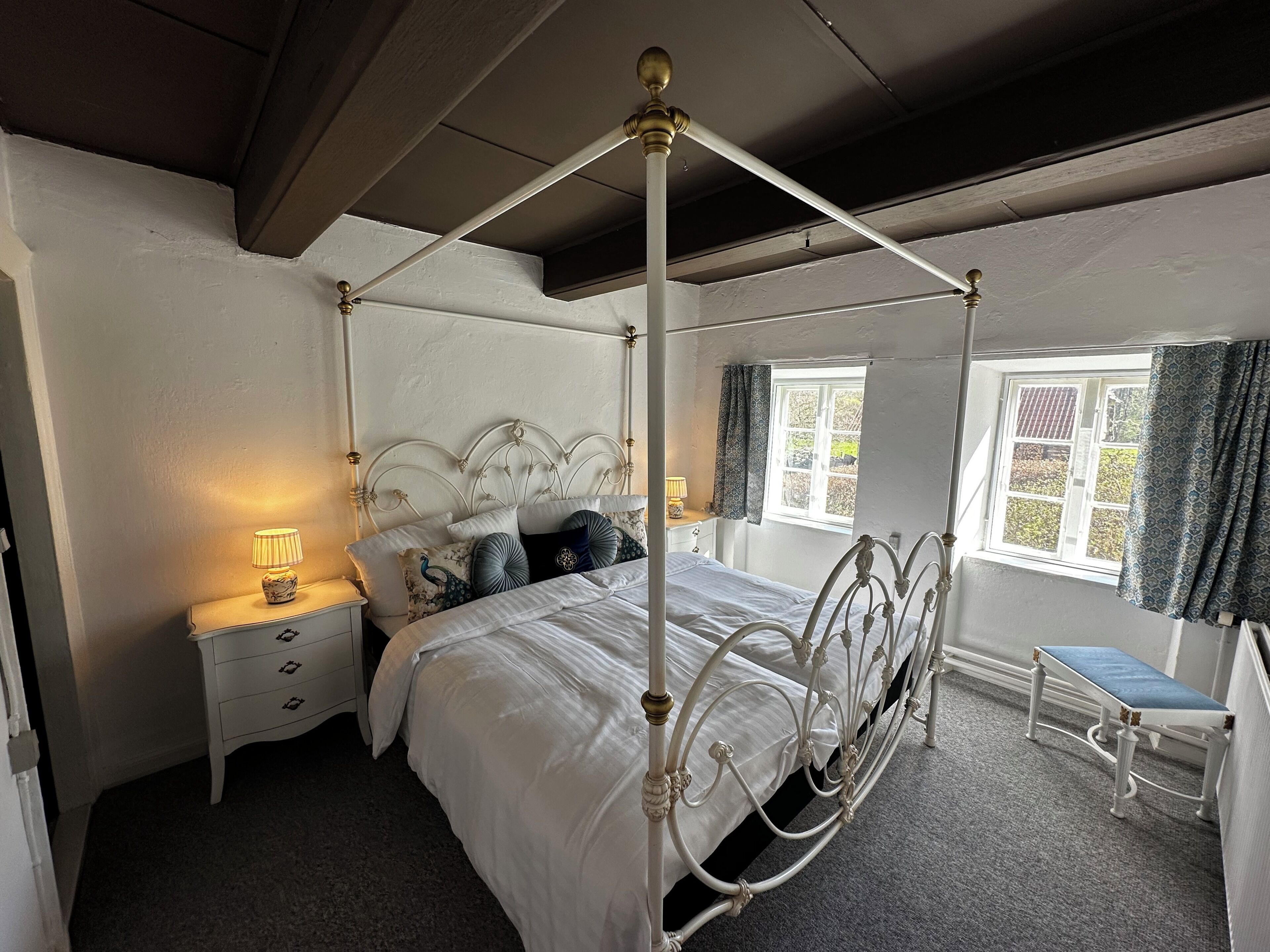 A four-poster bed with a canopy, a bedside table with a lamp, a small bench, and a window with curtains.