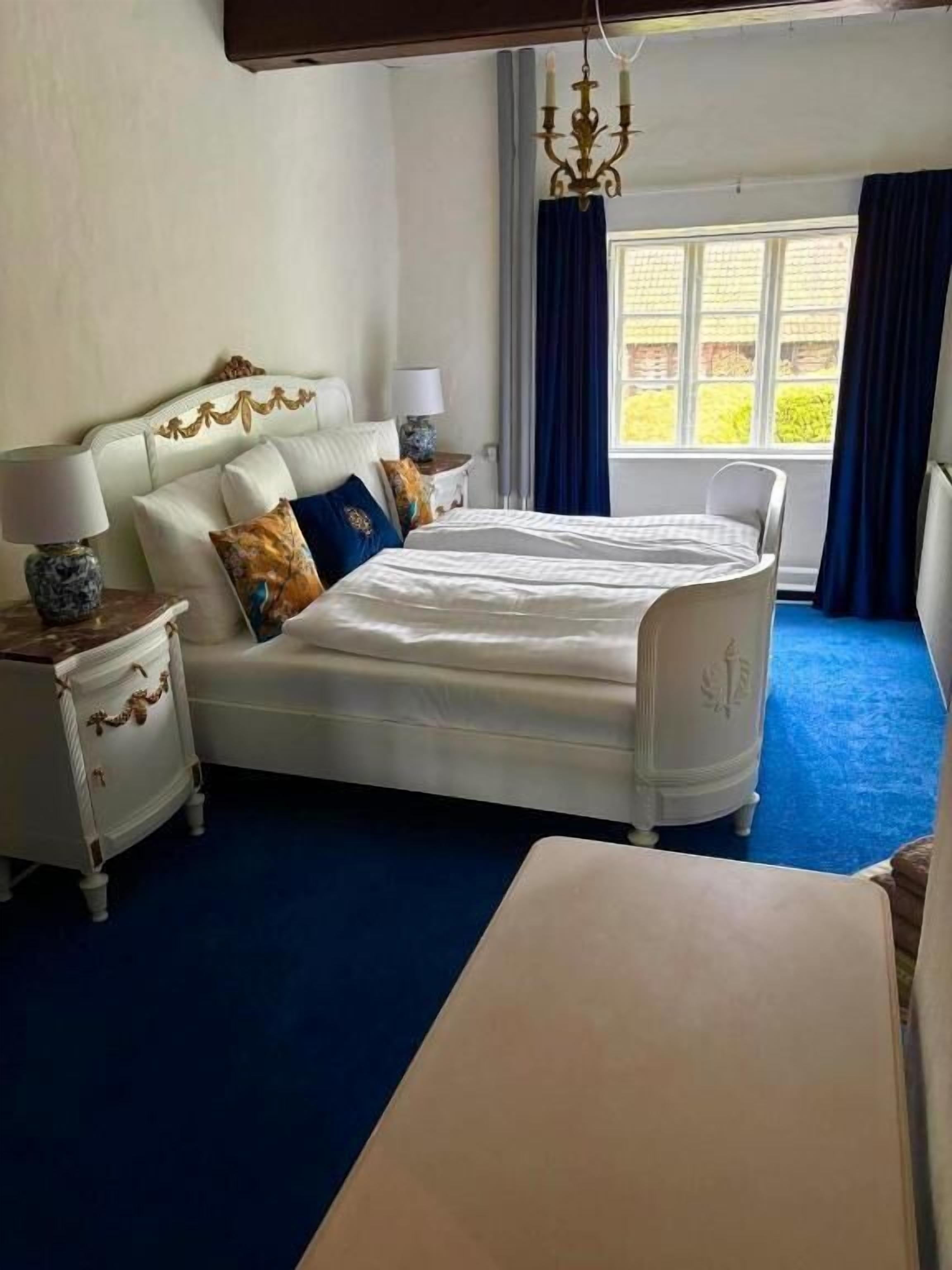 Une chambre à coucher avec un grand lit, deux tables de chevet avec des lampes, une fenêtre avec des rideaux et un lustre.