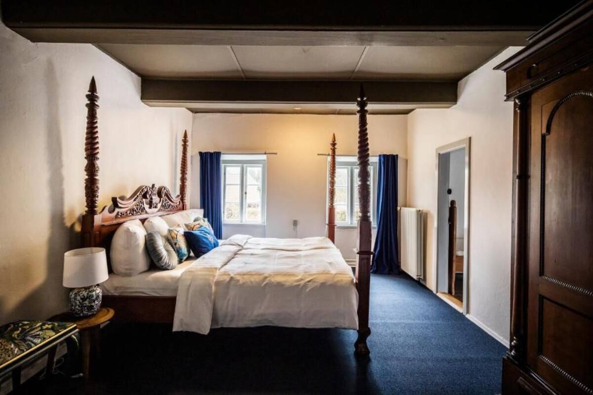 A four-poster bed with a wooden headboard, a bedside table with a lamp, and a window with curtains.