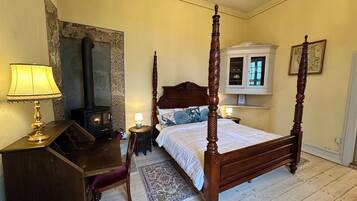 A traditional four-poster bed with a wooden headboard, a small bedside table with a lamp, a decorative rug, and a built-in wood-burning stove.