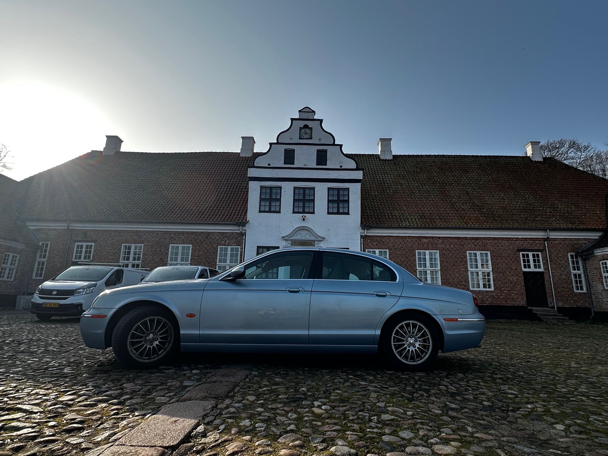 Ein Gebäude mit einem Glockenturm, parkenden Autos und einem Kopfsteinpflastergrund.