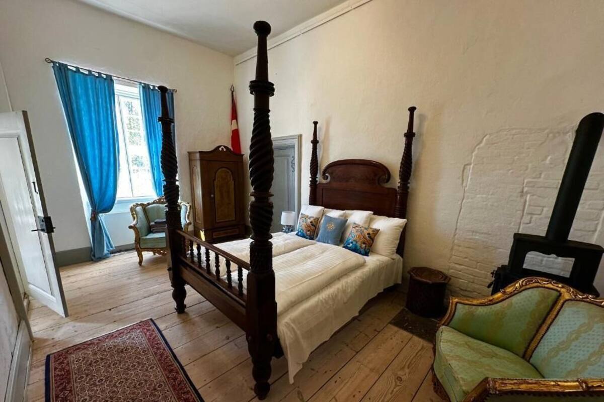 A four-poster bed with a wooden headboard, a green armchair, and a wooden chest of drawers.