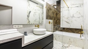 Ein modernes Badezimmer mit Marmorwaschtisch, einem runden Waschbecken, einer Duschkabine aus Glas und einem goldfarbenen Duschkopf.