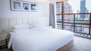 Quarto de hotel com uma cama grande, mesa de cabeceira, luminária na parede e janela com vista para a cidade.