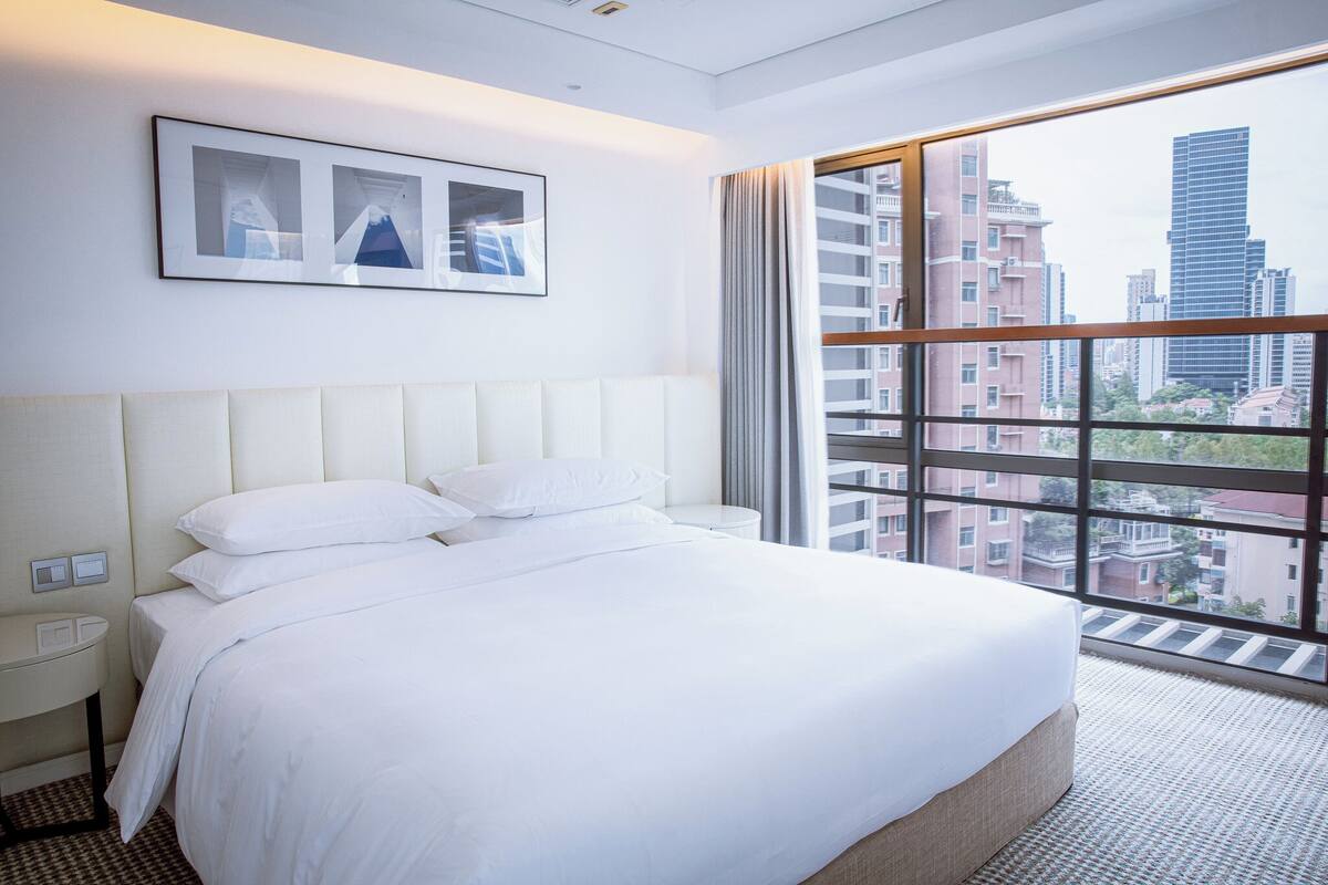 A hotel room with a large bed, a bedside table, a wall-mounted light, and a window with a city view.