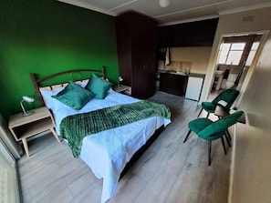 Une chambre à coucher avec un lit, des oreillers verts, une table de chevet avec une lampe, un fauteuil vert et une vue sur la cuisine et la salle de bain.