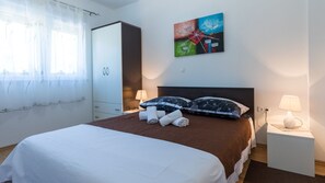 Ein Hotelzimmer mit Bett, zwei Nachttischlampen, einem Bild an der Wand, einem Kleiderschrank und einem Fenster mit durchsichtigen Vorhängen.