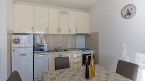 Eine kompakte Küche mit Kühlschrank, Mikrowelle, Kaffeemaschine und Herd. Ein Esstisch mit Stühlen und eine Uhr an der Wand sind vorhanden.