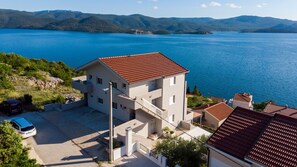 A multi-story building with a red-tiled roof, a parking area, and a view of a large body of water with hills in the background. - Apartman Arapovic 5 by Villas Guide (Klek)