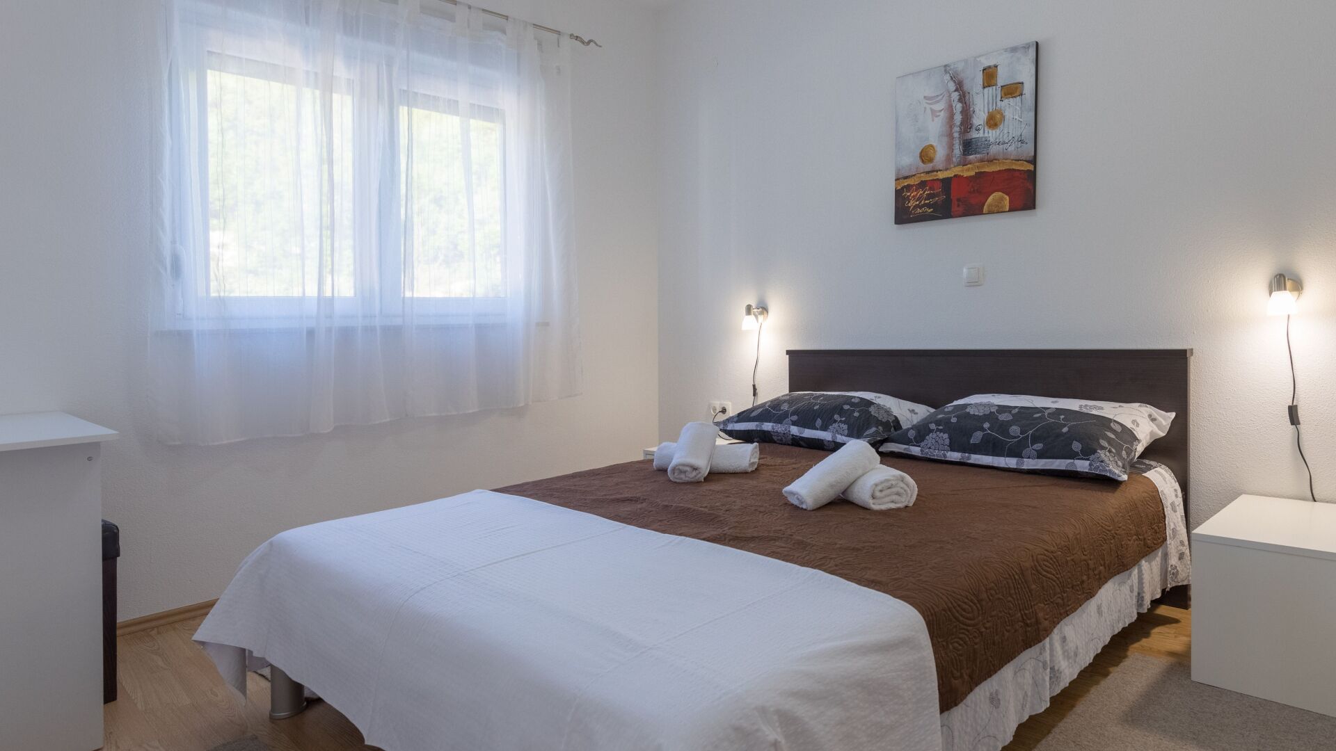 Una cama bien hecha con ropa blanca y un cabecero marrón, dos mesitas de noche fijadas a la pared con lámparas, una ventana con cortinas translúcidas y un cuadro en la pared.