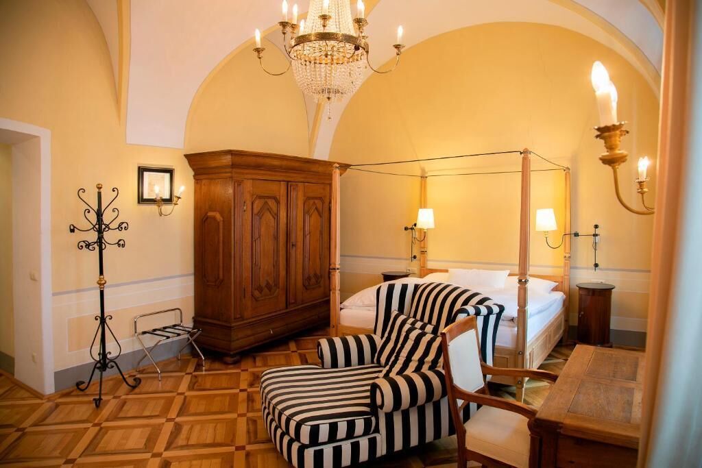 Ein Zimmer mit einem Bett, einem Holzkabinett, einem gestreiften Sessel, einer Chandelierleuchte und einer Wandlampe.