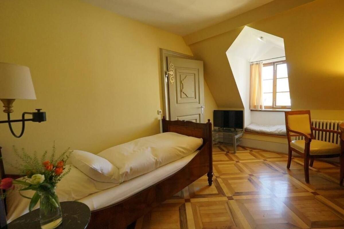 A single bed with a wooden frame, a bedside table with a lamp, a chair, a radiator, and a window with curtains.