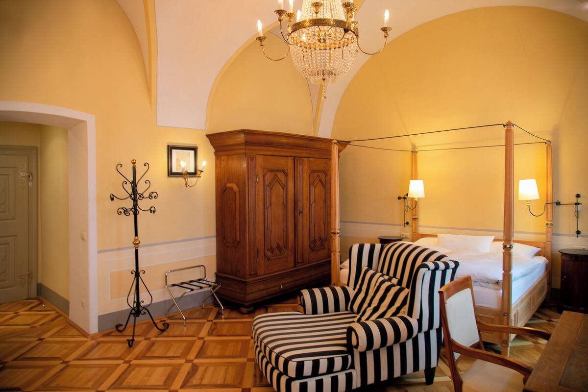 A bedroom with a four-poster bed, a wooden wardrobe, a striped armchair, and a standing lamp.