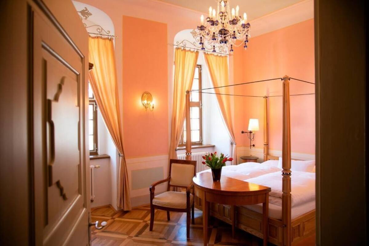 A room with a four-poster bed, a chandelier, a table with a vase, and a chair.