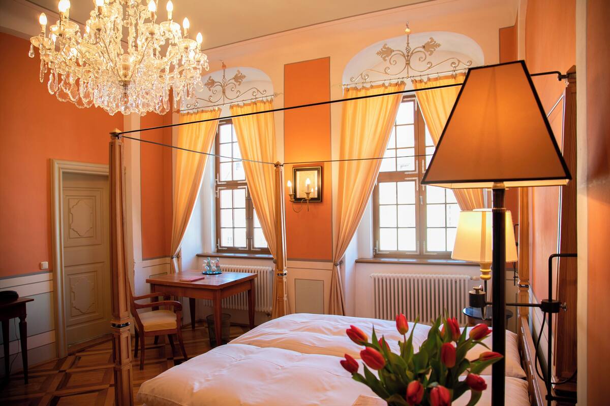 A hotel room with a bed, a desk, a chair, a chandelier, and a window with curtains.