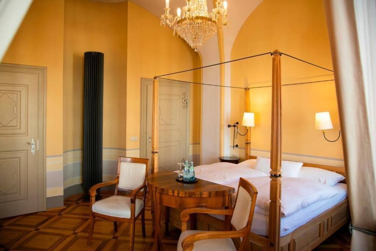 A four-poster bed, a chandelier, a wooden table with chairs, and a tall green vase.