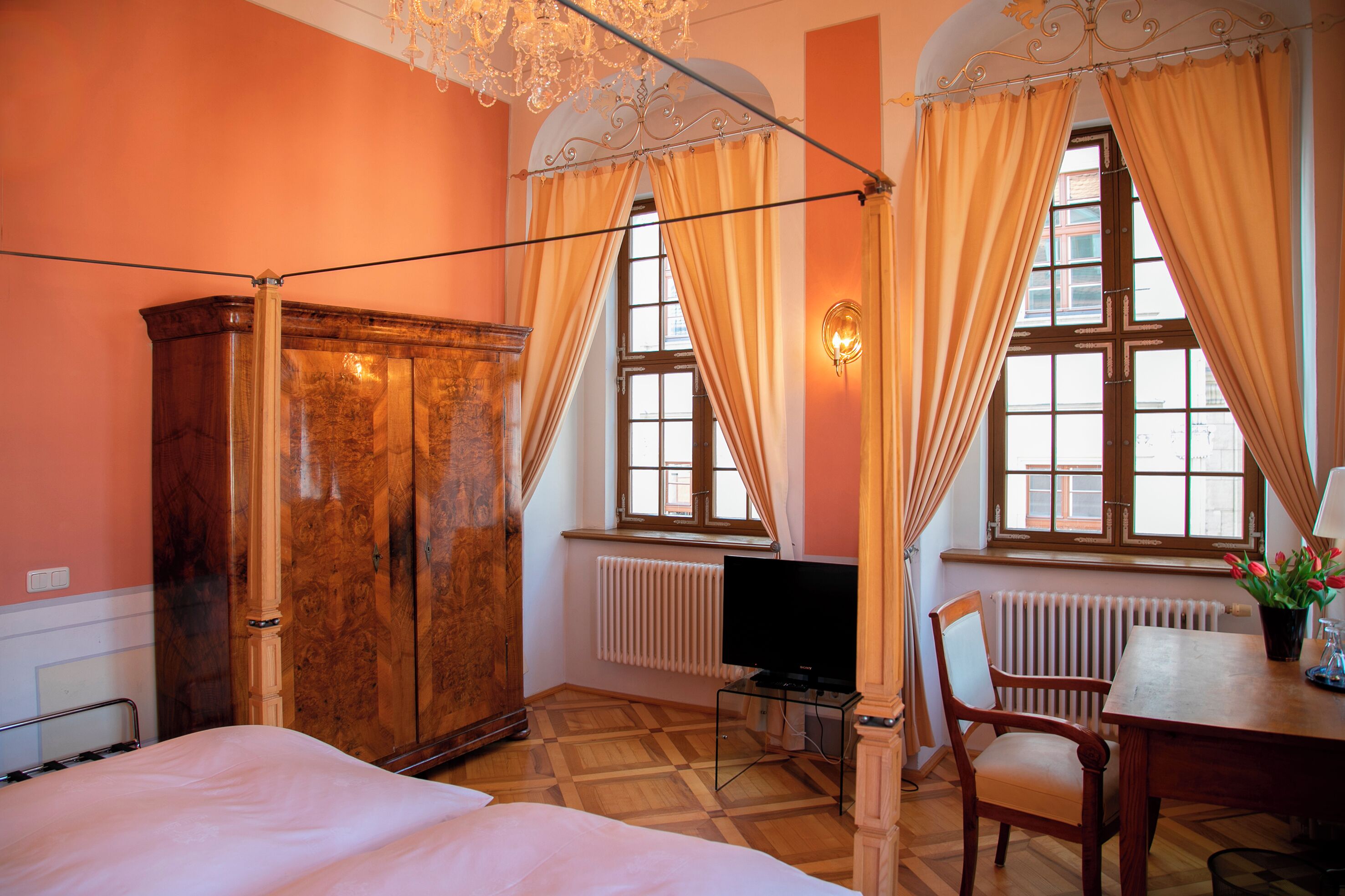 A room with a bed, a wooden wardrobe, a table with a chair, a television, and a window with curtains.
