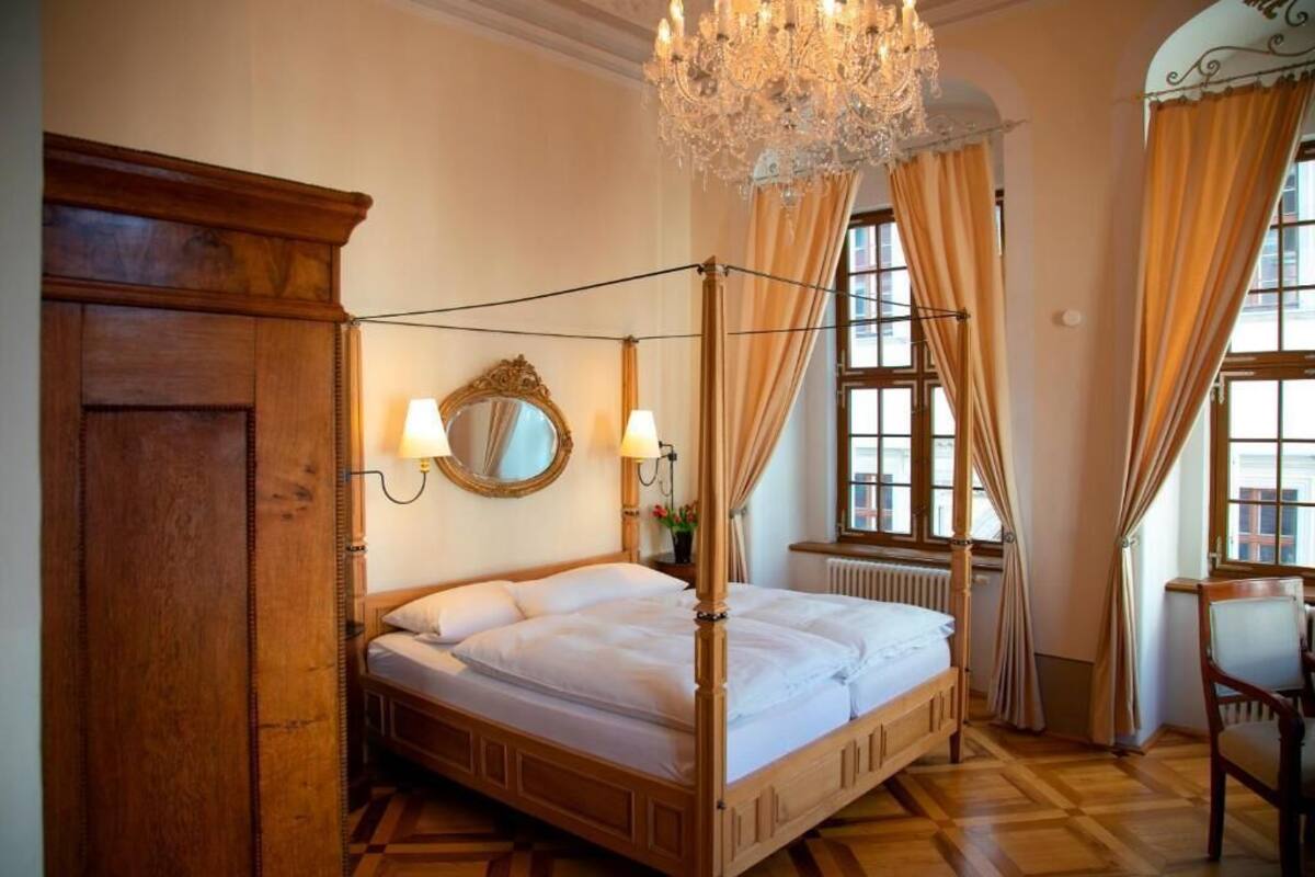 A four-poster bed with a mirror and wall sconces, a chandelier, and a view of the street through large windows.
