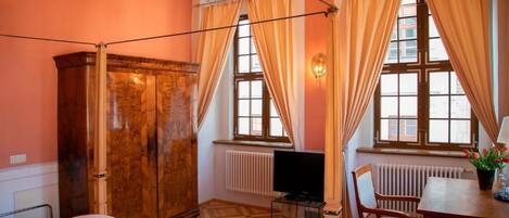 Una habitación con cama, un armario de madera, un escritorio con silla, televisión y ventana con cortinas.