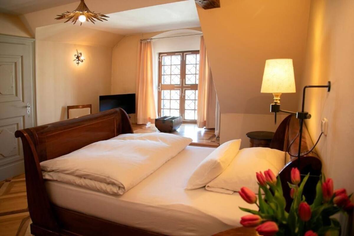 A double bed with white linens, a bedside lamp, a television, a window with curtains, and a vase with red flowers.