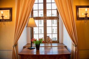 Ein Zimmer mit großem Fenster, Schreibtisch mit Lampe, Blumen in einer Vase und eine dekorative Pferdefigur.