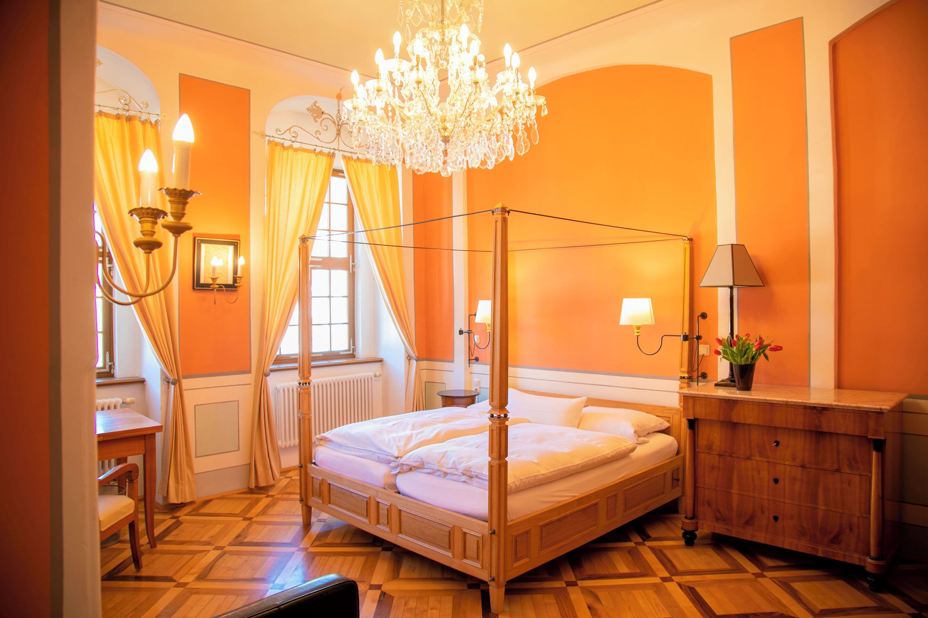 Una cama con dosel, una lámpara de araña, un aparador de madera y un escritorio con una silla en una habitación con paredes anaranjadas y suelo de madera.