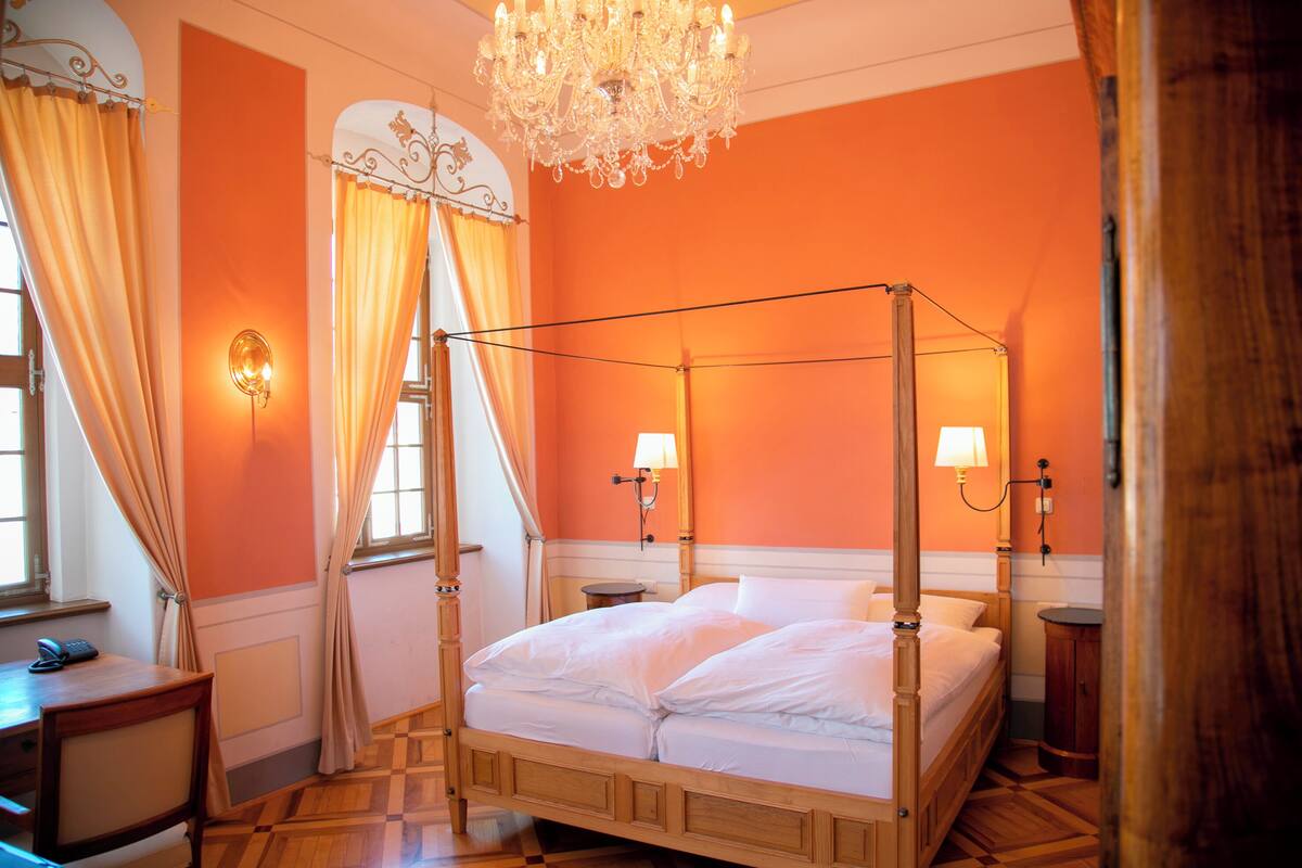 A four-poster bed with a wooden frame, two bedside tables, a chandelier, and a window with curtains.
