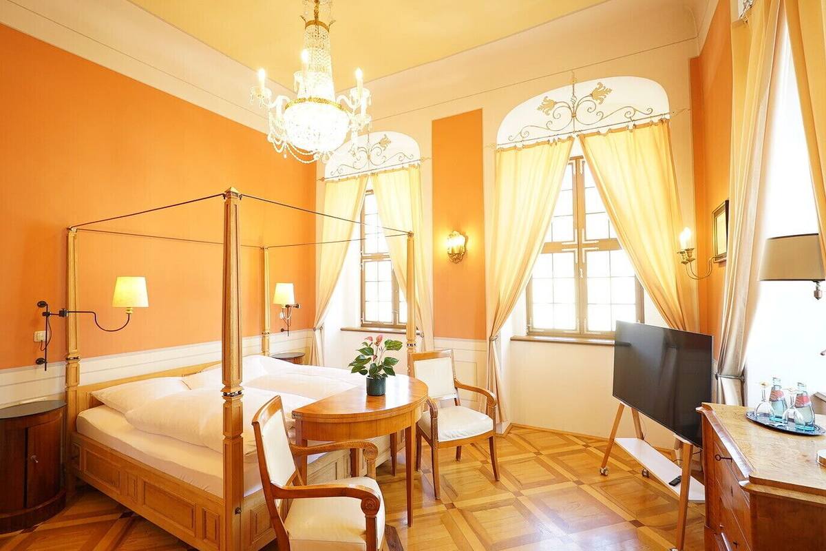 A four-poster bed, a television, a desk with a chair, and a potted plant in a room with orange walls and wooden flooring.