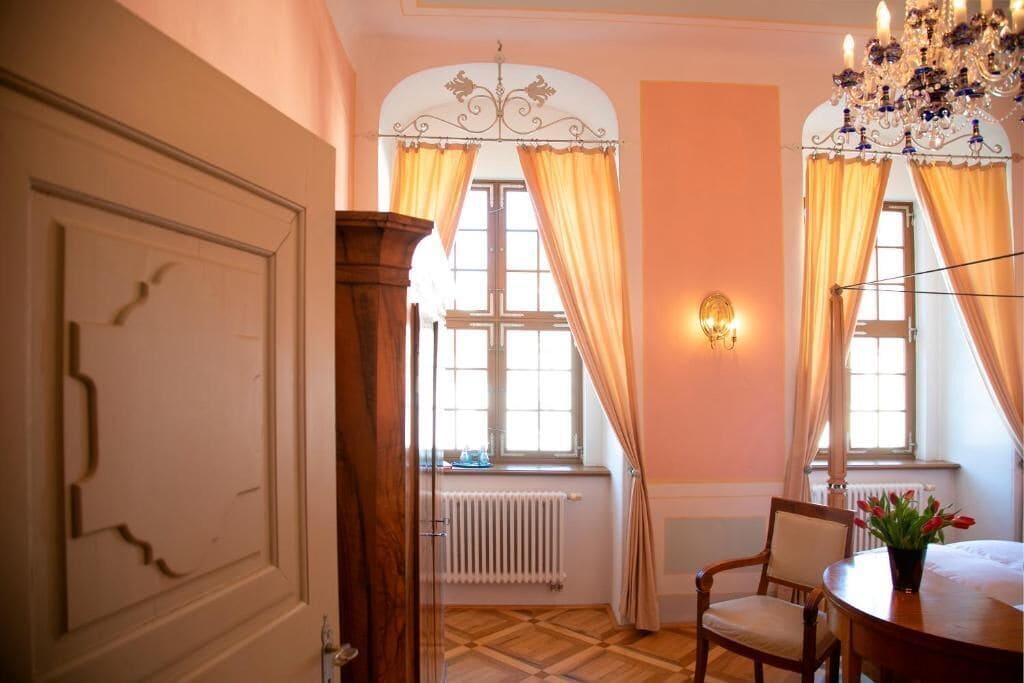 A room with a wooden door, a radiator, a chandelier, a table with a chair, and a window with curtains.