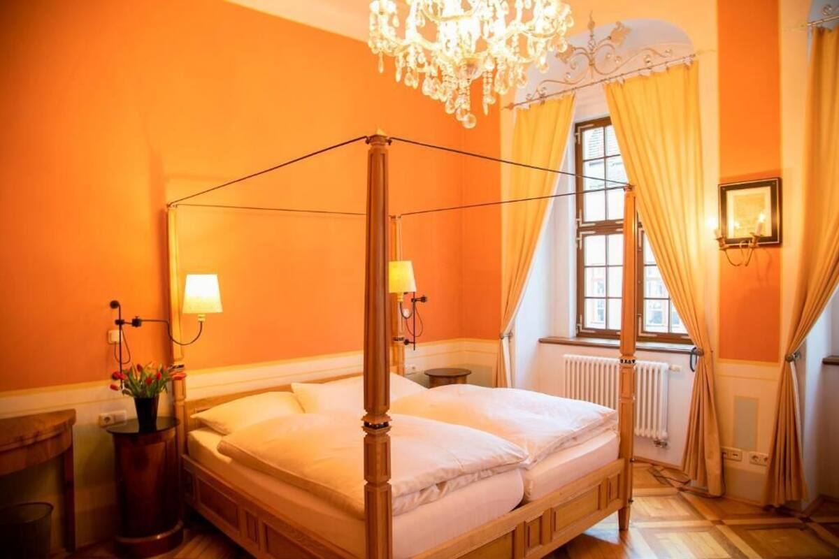 A four-poster bed in a room with orange walls, a chandelier, and a window with curtains.