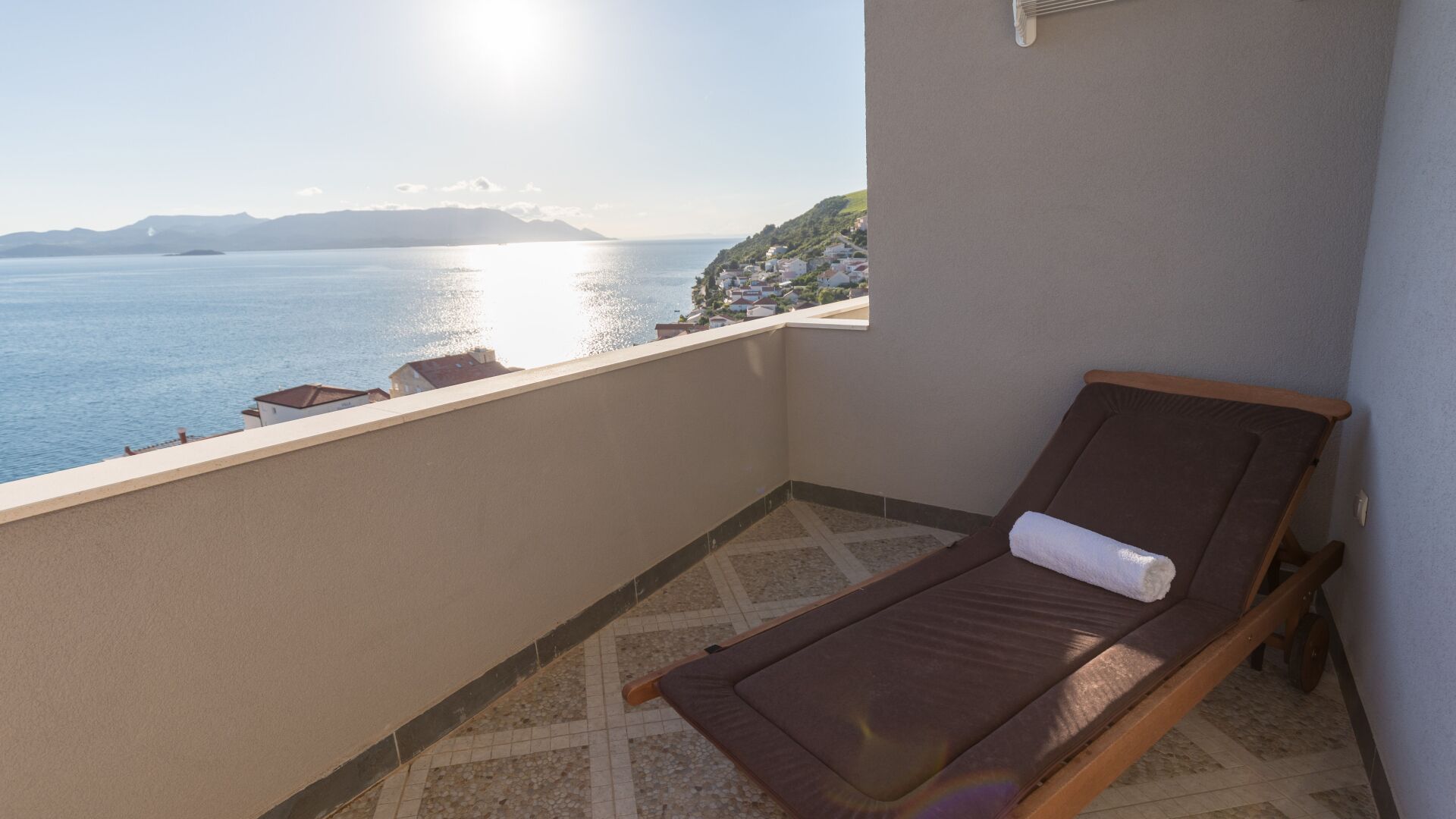 A balcony with a view of the sea, a sun lounger, and a tiled floor.