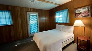 Un dormitorio con cama, una mesita de noche, una lámpara, una puerta con ventana y un cuadro en la pared.