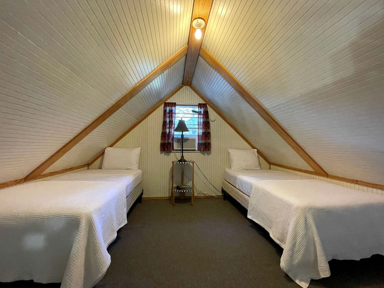 Um pequeno quarto sótão triangular com duas camas simples, uma janela com cortinas e uma luminária.