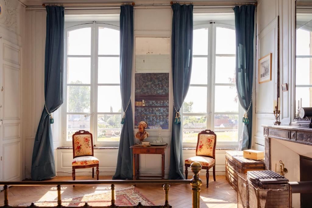 A room with large windows, two upholstered chairs, a small table with a bust, and a vintage typewriter.