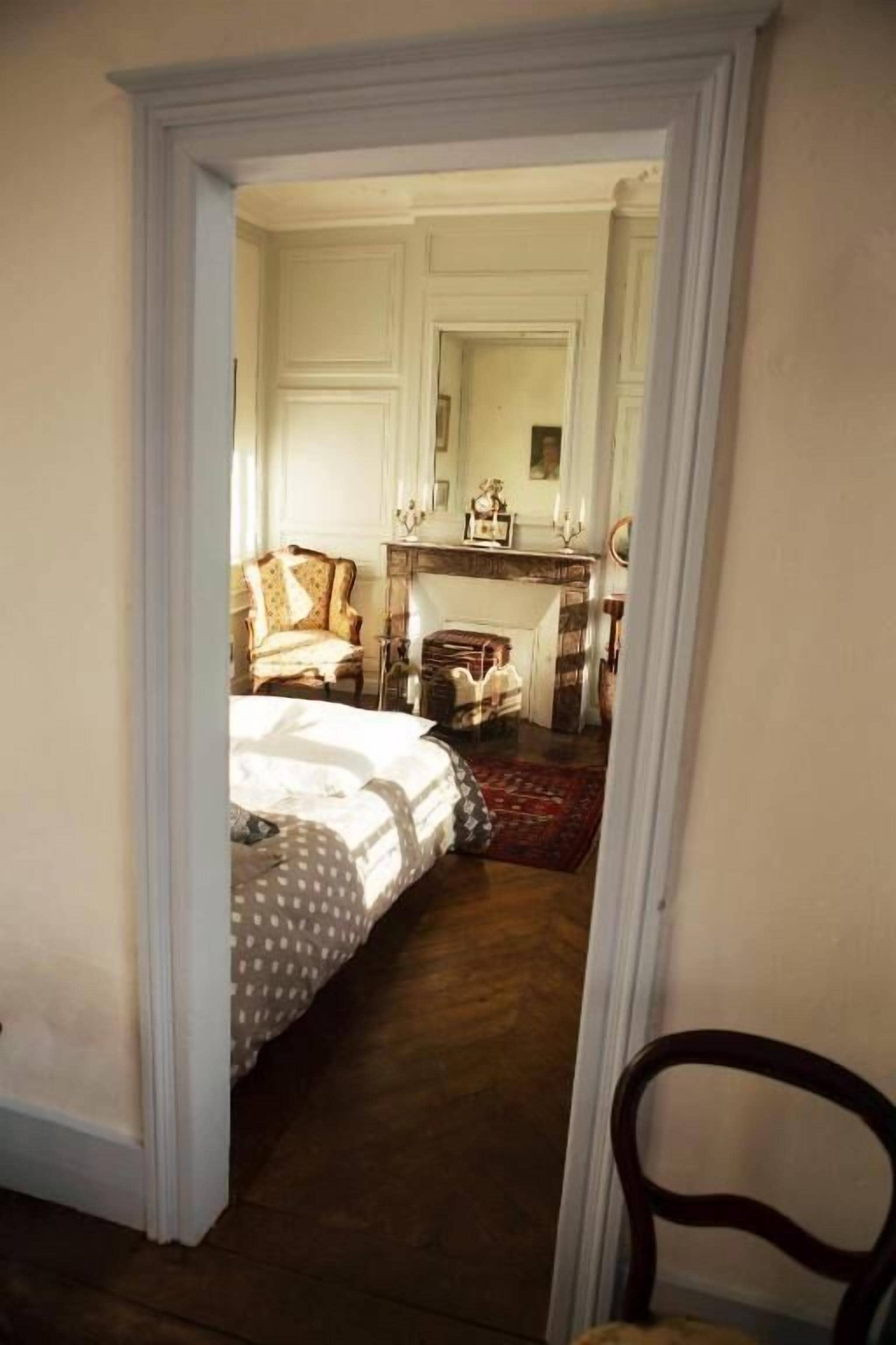 A narrow hallway leading to a bedroom with a bed, a chair, a small table, and a fireplace.