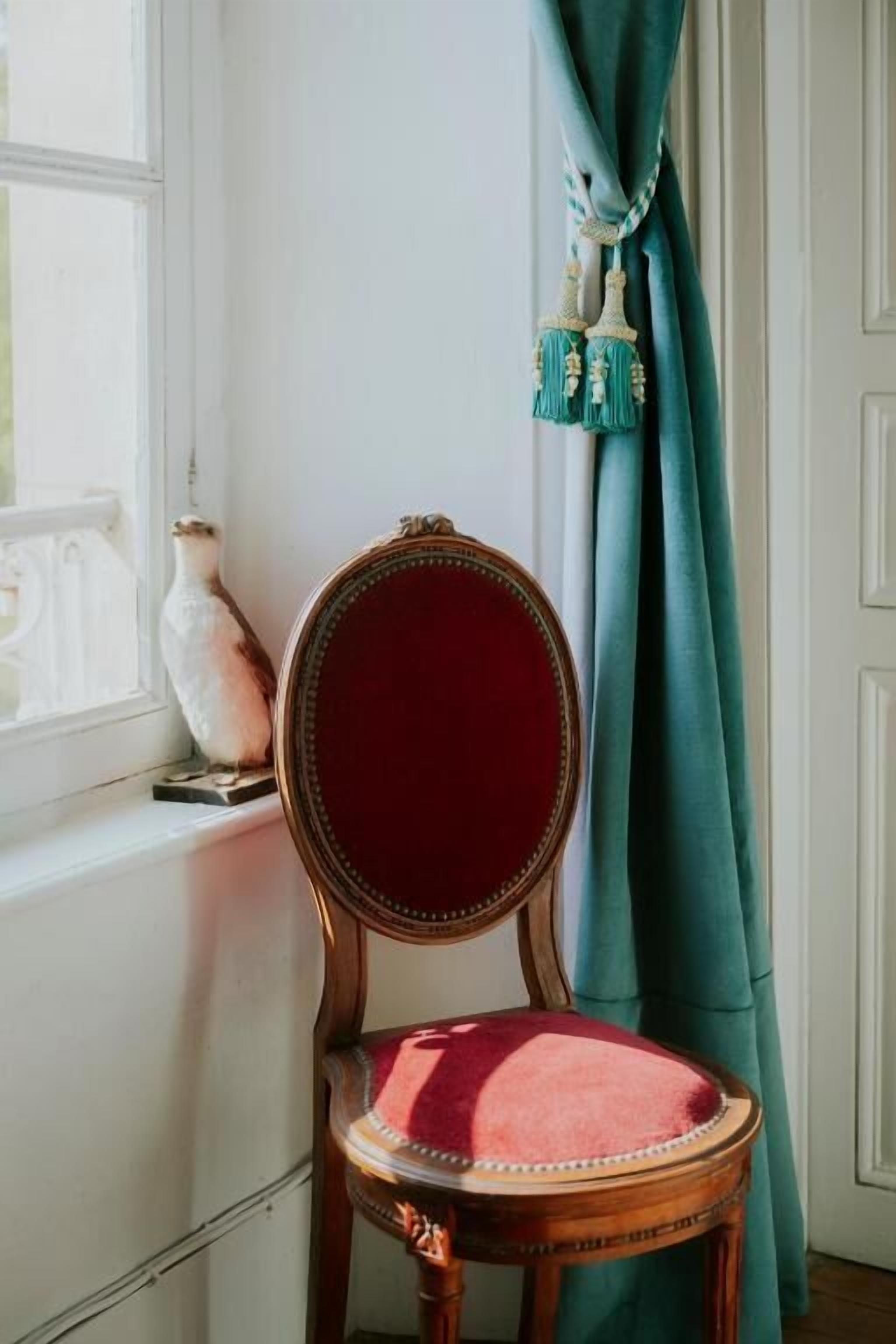Una silla con cojín rojo y estructura de madera, ubicada cerca de una ventana con molduras blancas.