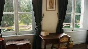 A room with a large window, a desk, a chair, a bed, and a small table.