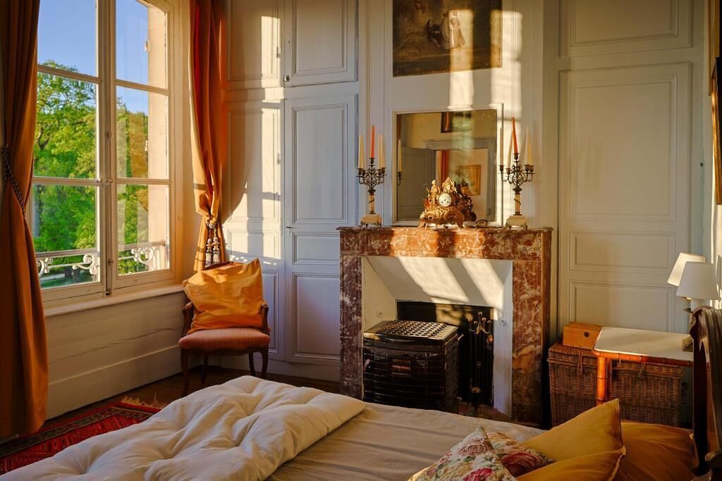 Un dormitorio con cama, chimenea, ventana con cortinas y vista a la vegetación.