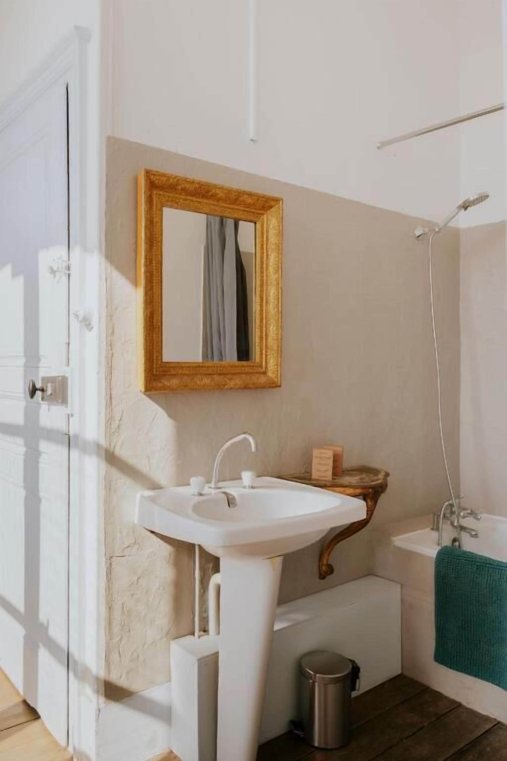 Een badkamer met een wit wastafelmeubel, een spiegel met gouden lijst, een bad en een handdoek aan de muur.