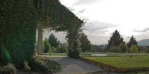 Un camí pavimentat que condueix a una zona coberta amb un arc de pedra, envoltada de vegetació i un parc amb un estany.