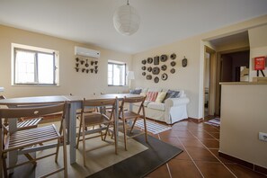 Zona pranzo con tavolo, sedie e divano. Si vede una finestra con tende, un condizionatore d'aria a parete e una parete decorata con piatti.