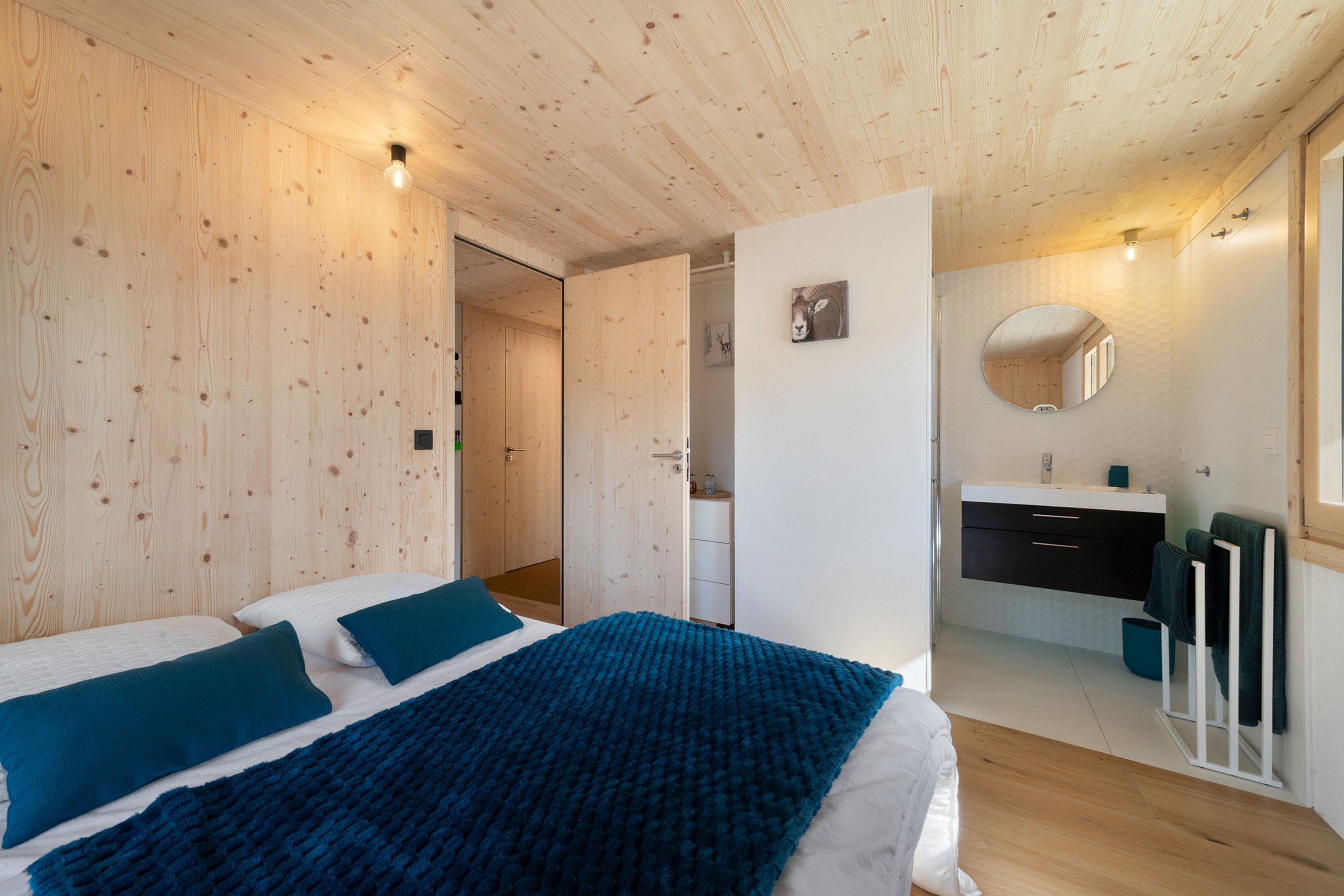 Une chambre avec un plafond en bois, un lit avec une literie bleue, une table de chevet et un miroir fixé au mur.