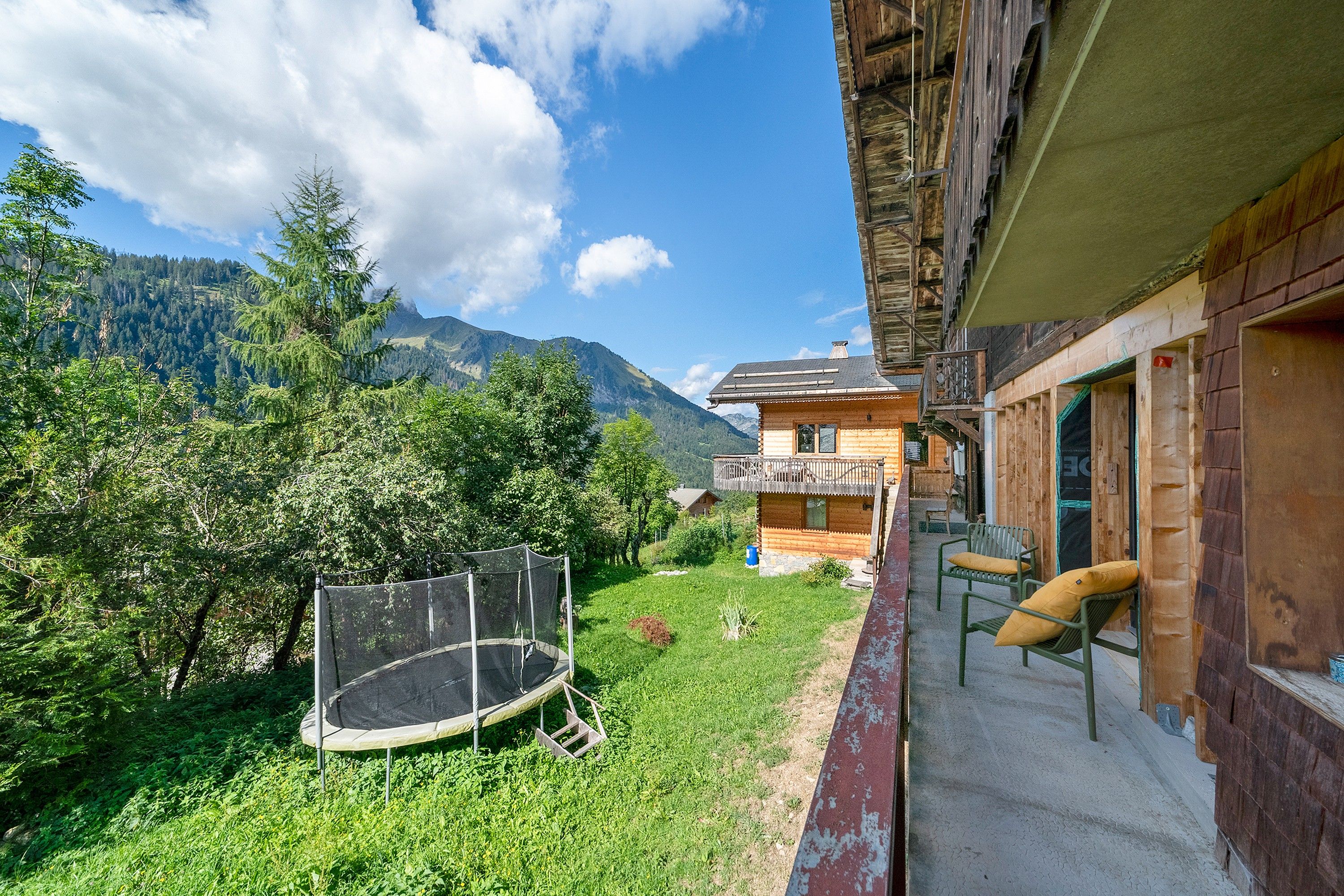 A wooden chalet with a balcony overlooking a grassy yard and a mountain backdrop.