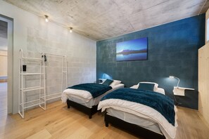 A bedroom with two beds, a white metal shelf unit, a painting of a landscape, and a blue accent wall. - Apartment 'C’Emile - 74390' with Mountain View, Shared Garden, and Wi-Fi (Châtel)