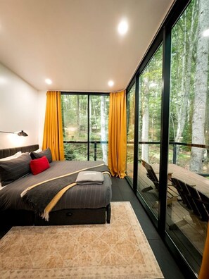 A bedroom with a large bed, a nightstand, a lamp, and a view of a forest through large windows. - Creekside Glass Suite - Unit 2 (Pet Friendly) (Mars Hill)