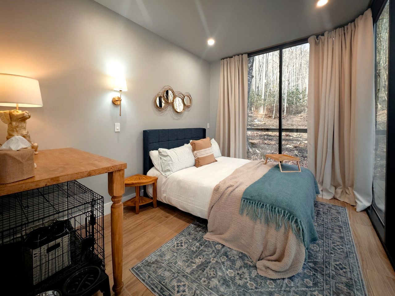 Een slaapkamer met een bed, nachtkastje, lamp en uitzicht op bomen.