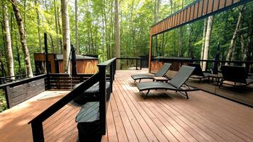 A wooden deck with lounge chairs, a bench, and a railing overlooking a forest.